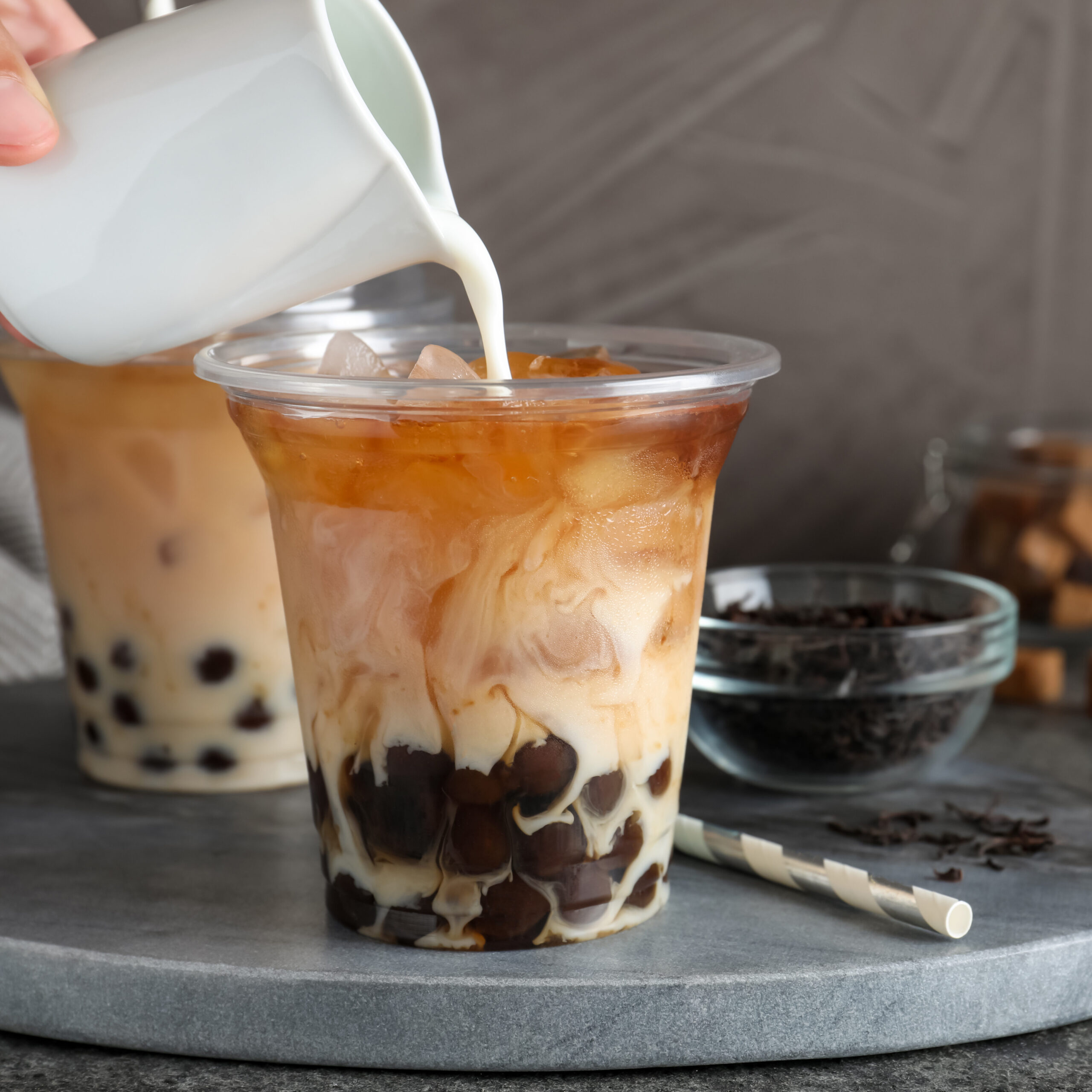 Person pouring milk into plastic cup with bubble tea at grey table, closeup